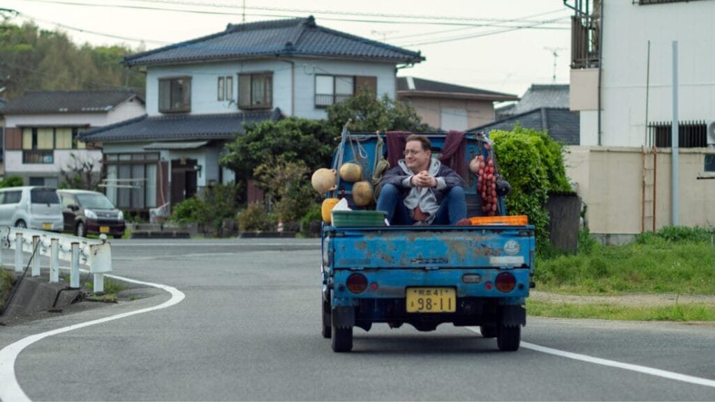 Brendan Fraser rides on the back of a comically small blue pickup truck across a suburban neighborhood in Tokyo, Japan in the feel-good comedy film RENTAL FAMILY.