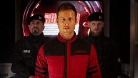 A furious Glen Powell wears a red uniform with the last name "Richards" printed on the chest as two armed guards walk him onto the Running Man game show stage on live television.