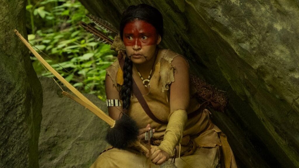 A female Native American warrior from the fictional Shokopiwah tribe holds up a bow and arrow with her face covered in red paint in episode 4 of HBO's IT: WELCOME TO DERRY.