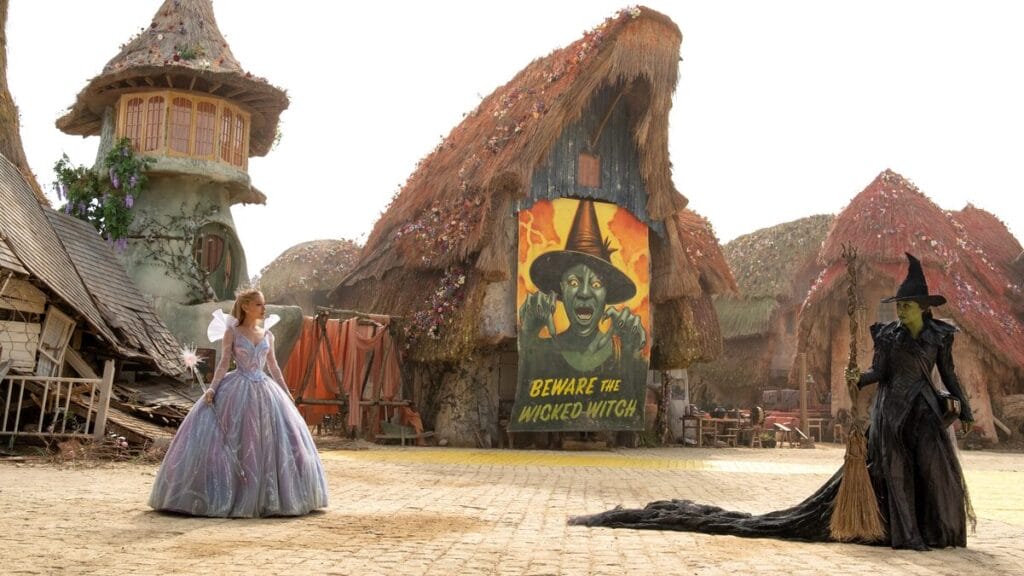 Ariana Grande as Glinda the Good and Cynthia Erivo and Elphaba Thropp face off in the middle of a village in Munchkinland while a poster depicting the evil Wicked Witch of the West hangs in between them in WICKED: FOR GOOD.