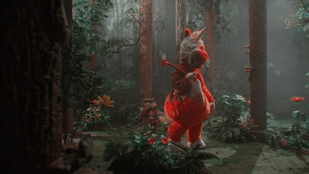 A killer in a huge red and fluffy unicorn costume holds up an axe in the new horror films BUDDY, premiering at the 2026 Sundance Film Festival. 