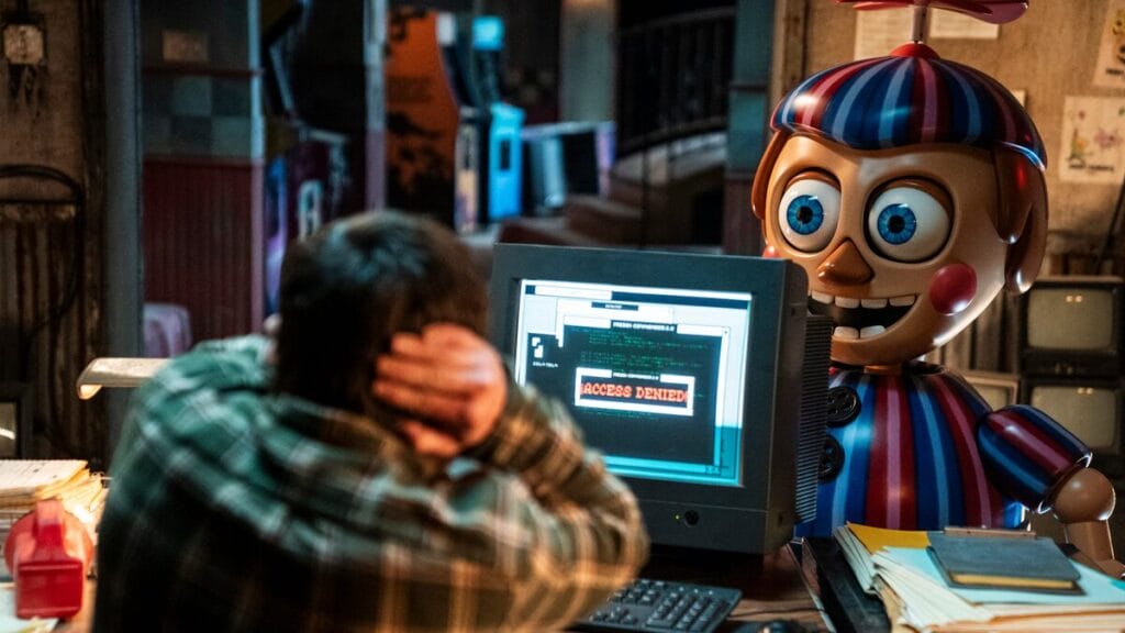 Josh Hutcherson stars as Mike Schmidt sitting at the security desk of Freddy's Pizzeria and covering his ears in pain as he's tormented by a large animatronic of Balloon Boy standing right in front of him in the horror sequel FIVE NIGHTS AT FREDDY'S 2. 