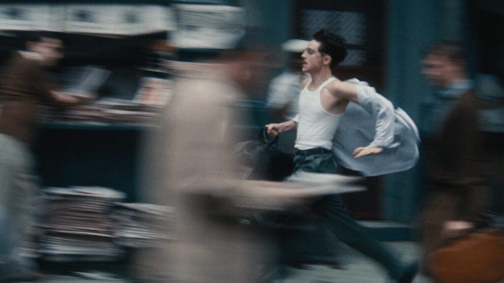 A blurry shot of Timothée Chalamet running down a busy street in 1950s New York City with his shirt almost flying off in the A24 film MARTY SUPREME. 