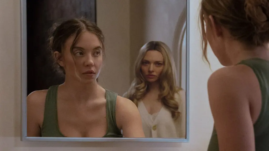 Sydney Sweeney looks up into a large bathroom mirror while cleaning a sink only to find a crazy-looking Amanda Seyfried standing right behind her in the psychological thriller THE HOUSEMAID.