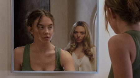 Sydney Sweeney looks up into a large bathroom mirror while cleaning a sink only to find a crazy-looking Amanda Seyfried standing right behind her in the psychological thriller THE HOUSEMAID.