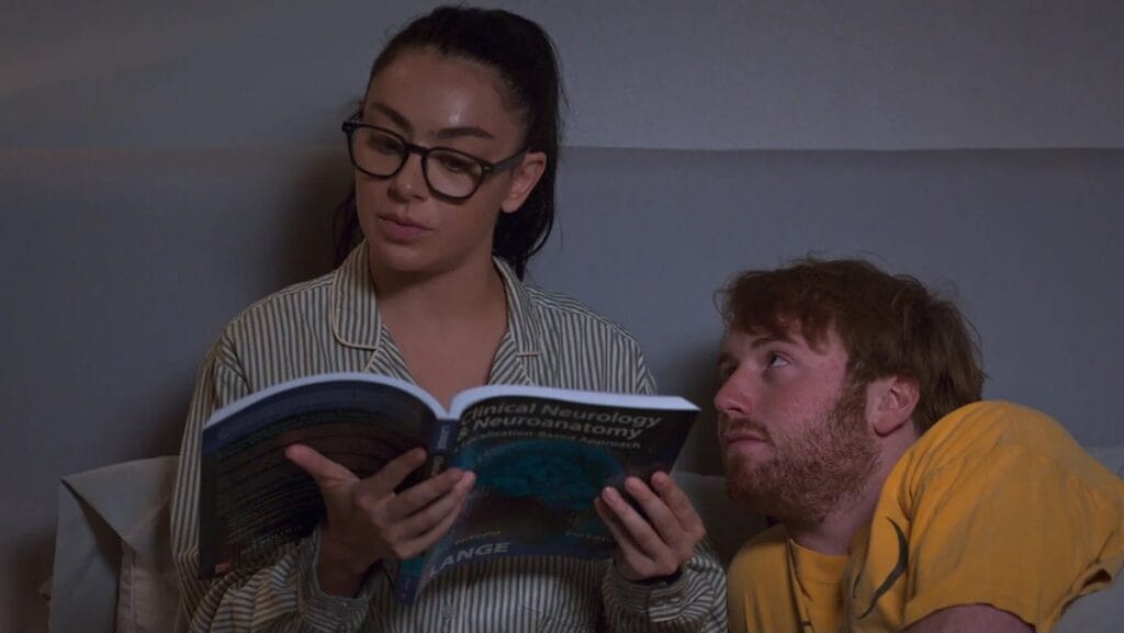 Charli xcx reads studiously in bed with reading glasses while Cooper Hoffman cuddles pathetically next to her in the erotic comedy I WANT YOUR SEX.