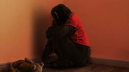 Ben the killer chimpanzee, wearing an orange shirt, sits creepily in the corner of his room with an old teddy bear lying next to him on the floor on the official poster for the horror movie PRIMATE.