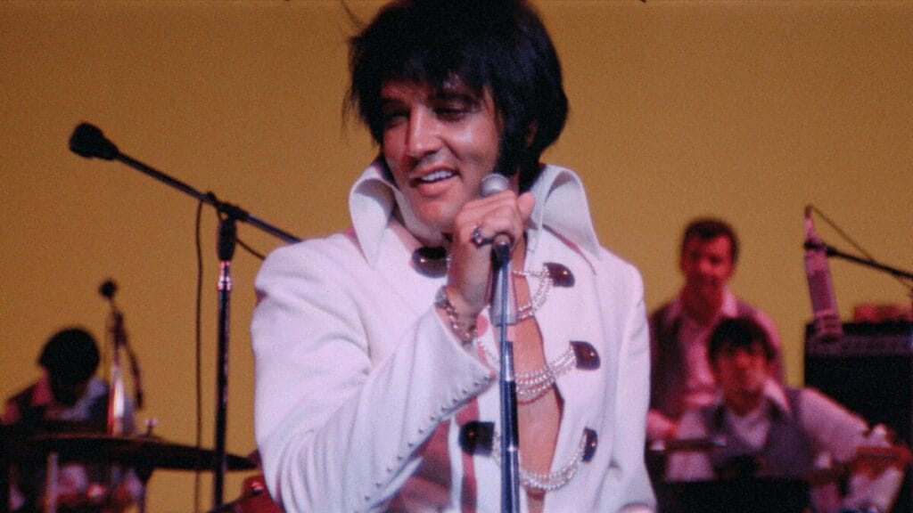 A close-up shot of Elvis Presley giving a huge smile while performing on stage in his iconic white leather jump suit from the EPiC concert film.