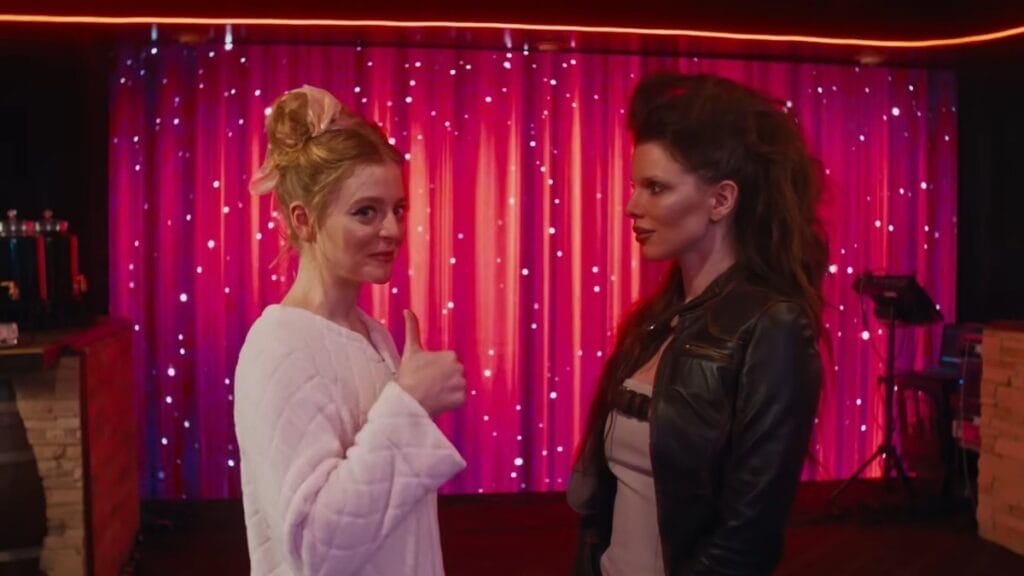 Anna Baryshnikov looks to the camera and gives a thumbs up while being interviewed by Julia Fox as part of a fashion competition show in the satirical comedy IDIOTKA. 
