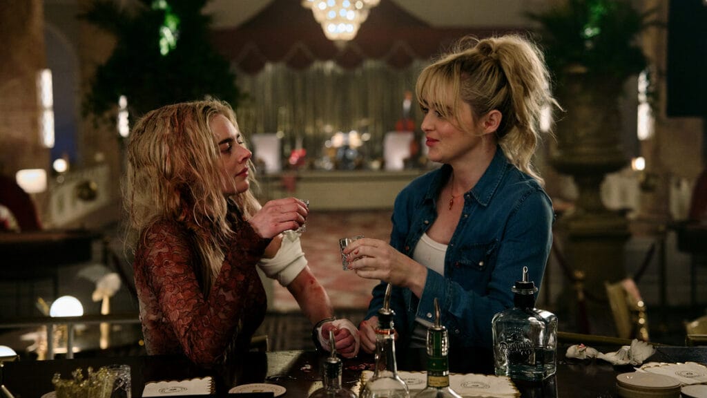 Samara Weaving wearing a blood-soaked wedding dress and Kathryn Newton wearing a torn jean shirt hold up two shot glasses as they give a toast while sitting together at an empty luxurious bar in READY OR NOT 2.