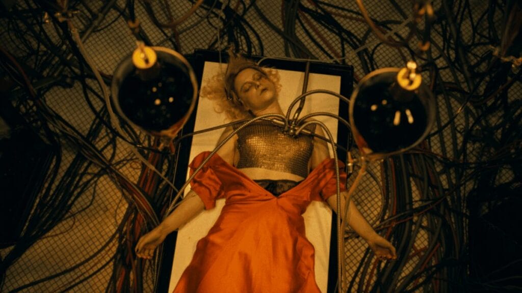 The cold corpse of Jessie Buckley in a bright orange dress lies still on a creepy operating table right before she's brought to life as the Bride of Frankenstein.