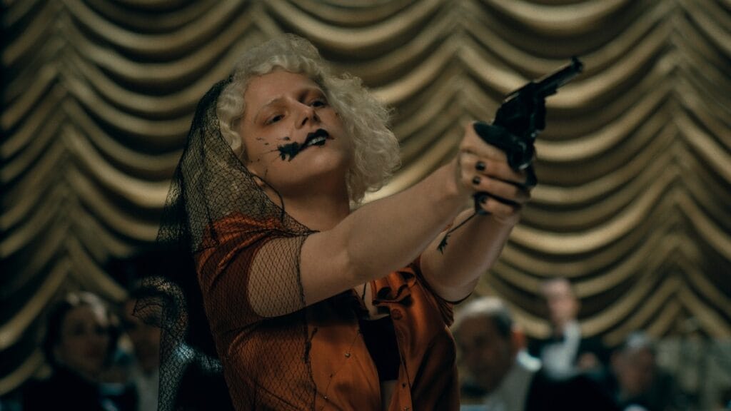 Jessie Buckley stars as the Bride of Frankenstein aiming a pistol in the air as she holds a bunch of people in a fancy ballroom hostage in Maggie Gyllenhaal's THE BRIDE!