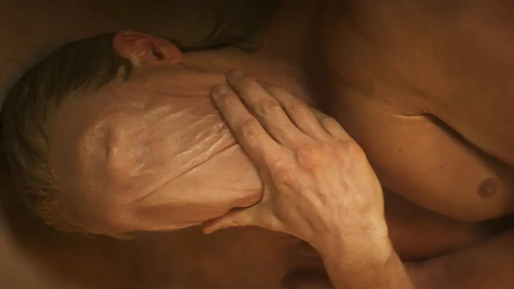 A horrific shot of Tom Rhys Harries as actor Matt Hagen wiping his eyes, nose, and mouth clear from his melted face while laying in a bathtub in the DC body horror movie CLAYFACE. 