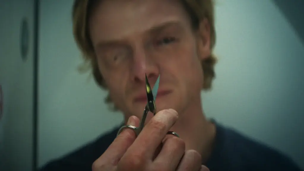 Tom Rhys Harries stars as actor Matt Hagen holding up a small pair of scissors to the camera as he prepares to cut his melting eyes open in a small airplane bathroom in the DC horror film CLAYFACE. 